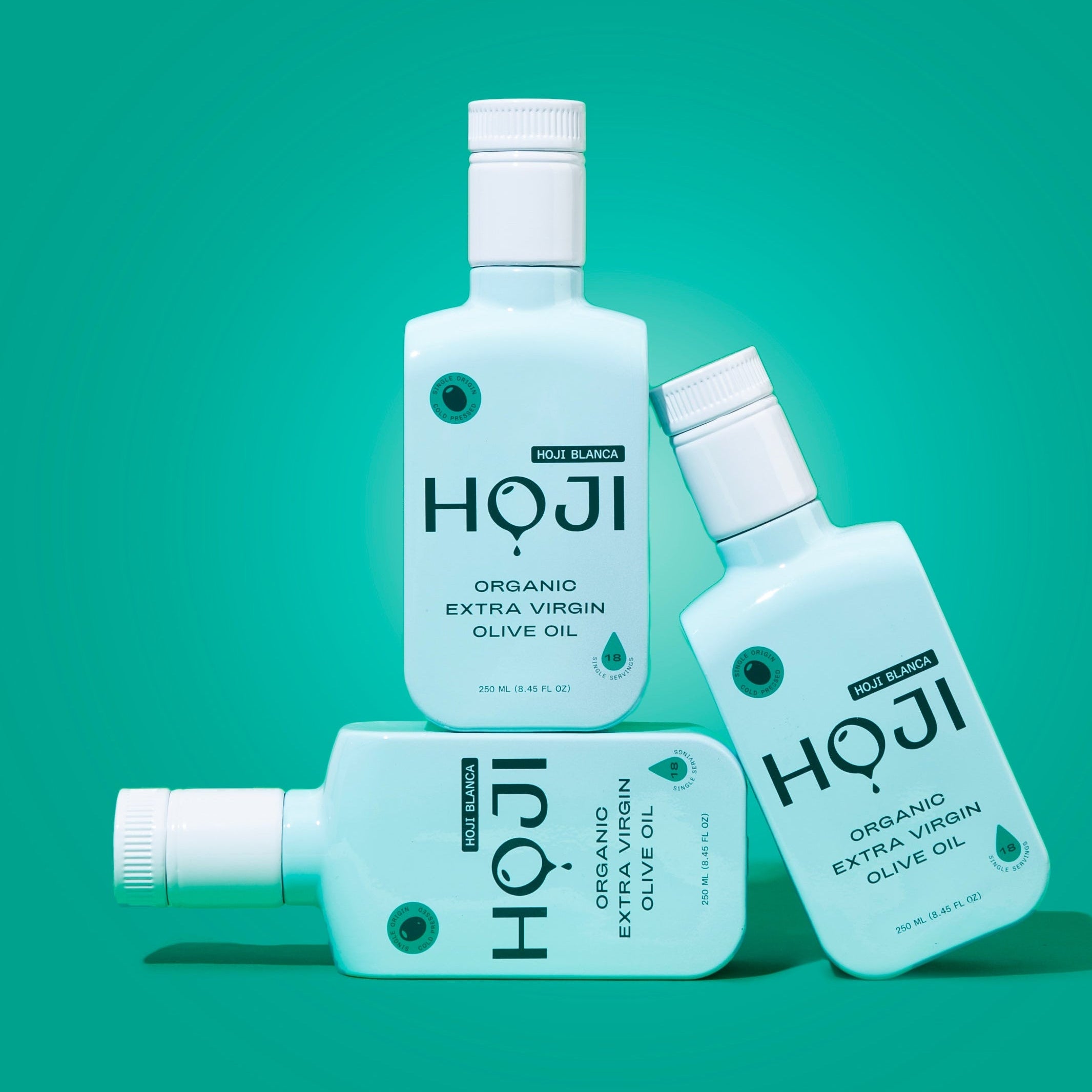 Three turquoise bottles of THE BOTTLE by Hoji are arranged on a bright teal background, with two upright and one lying horizontally in front.