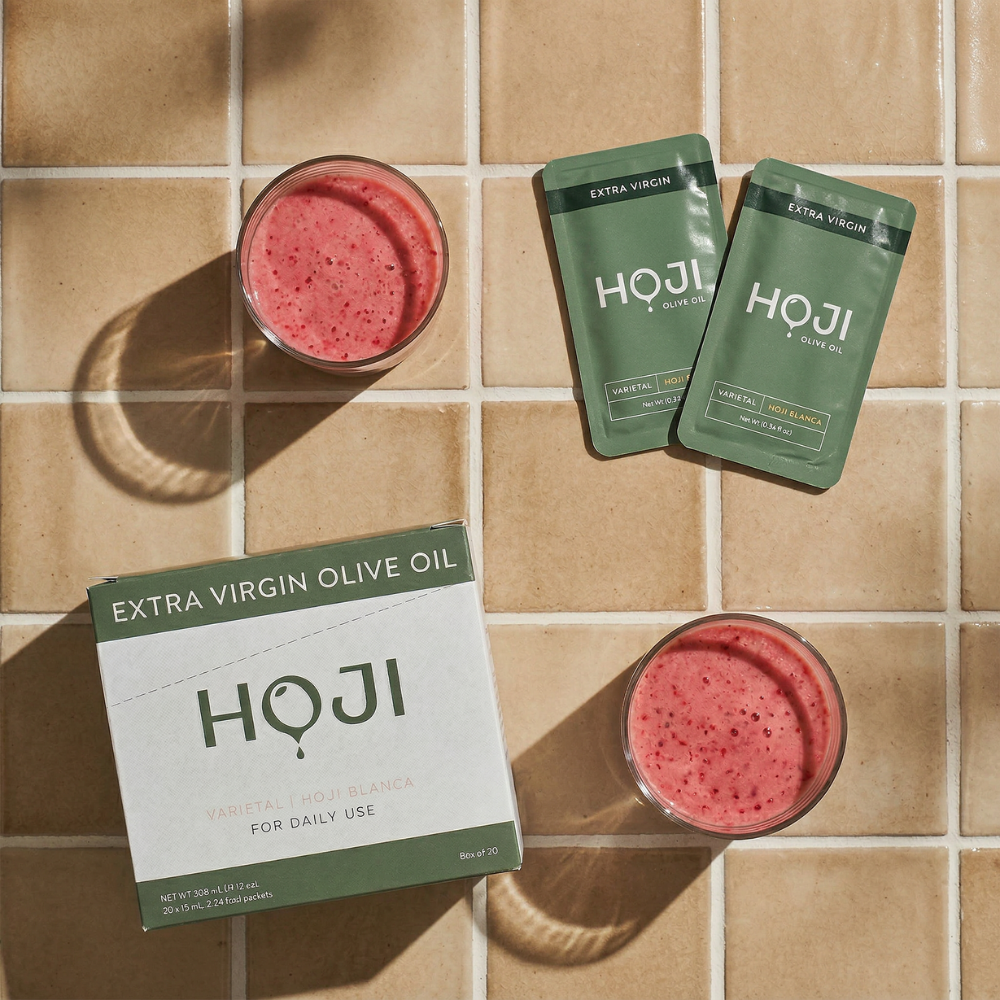 A box labeled "The 30 Pack" by My Store, two green single-serve olive oil packets, and two glasses of pink smoothie are arranged on beige tiles in bright sunlight.