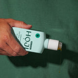 A hand pours a thin stream of oil from a light blue HOJI BLANCA BOTTLE by Hoji against a green background.
