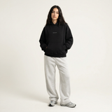 A person with long dark hair stands against a plain light background, wearing the oversized HOJI HOODIE by Hoji, light gray sweatpants, and gray sneakers, hands in the hoodie pocket with a neutral expression.