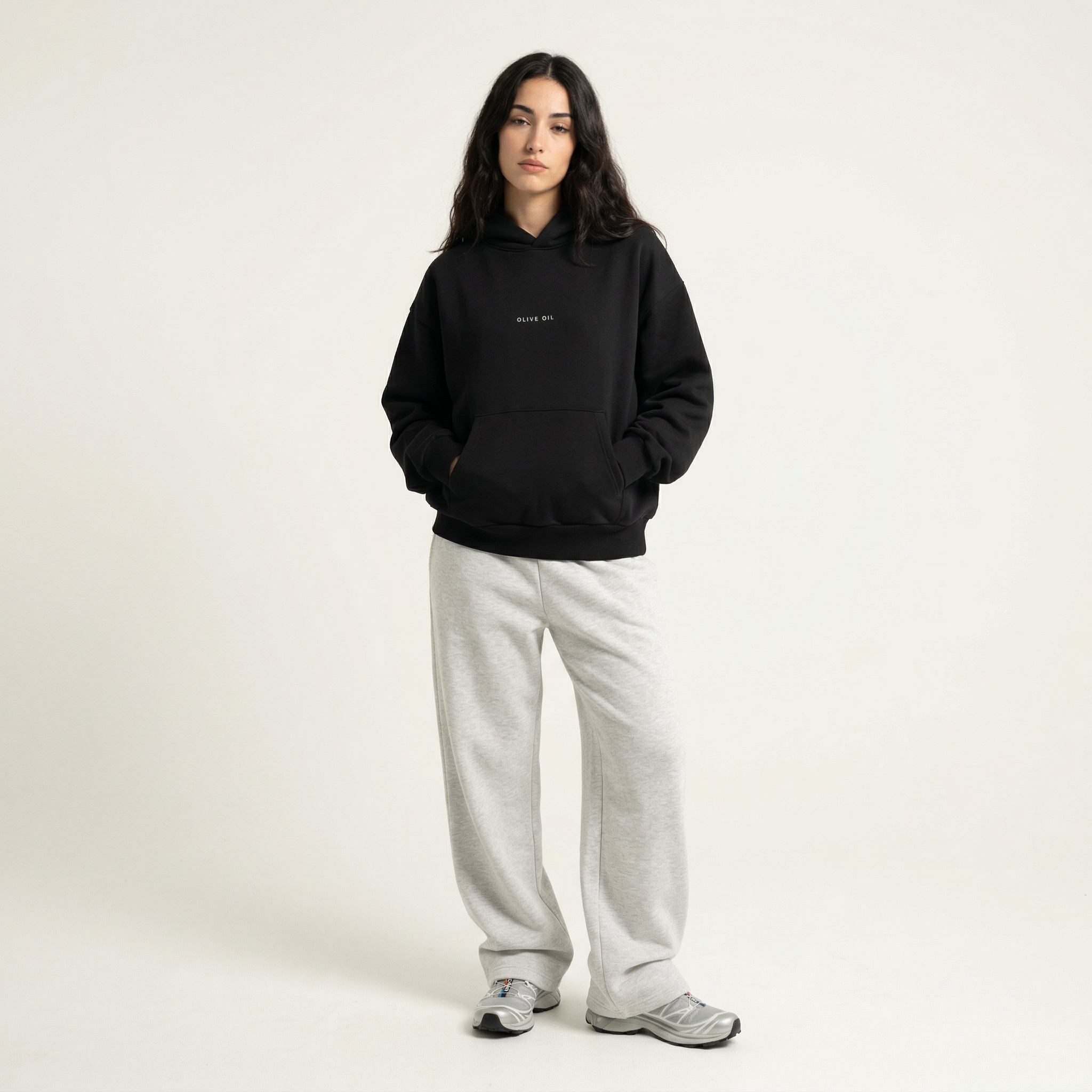 A person with long dark hair stands against a plain light background, wearing the oversized HOJI HOODIE by Hoji, light gray sweatpants, and gray sneakers, hands in the hoodie pocket with a neutral expression.