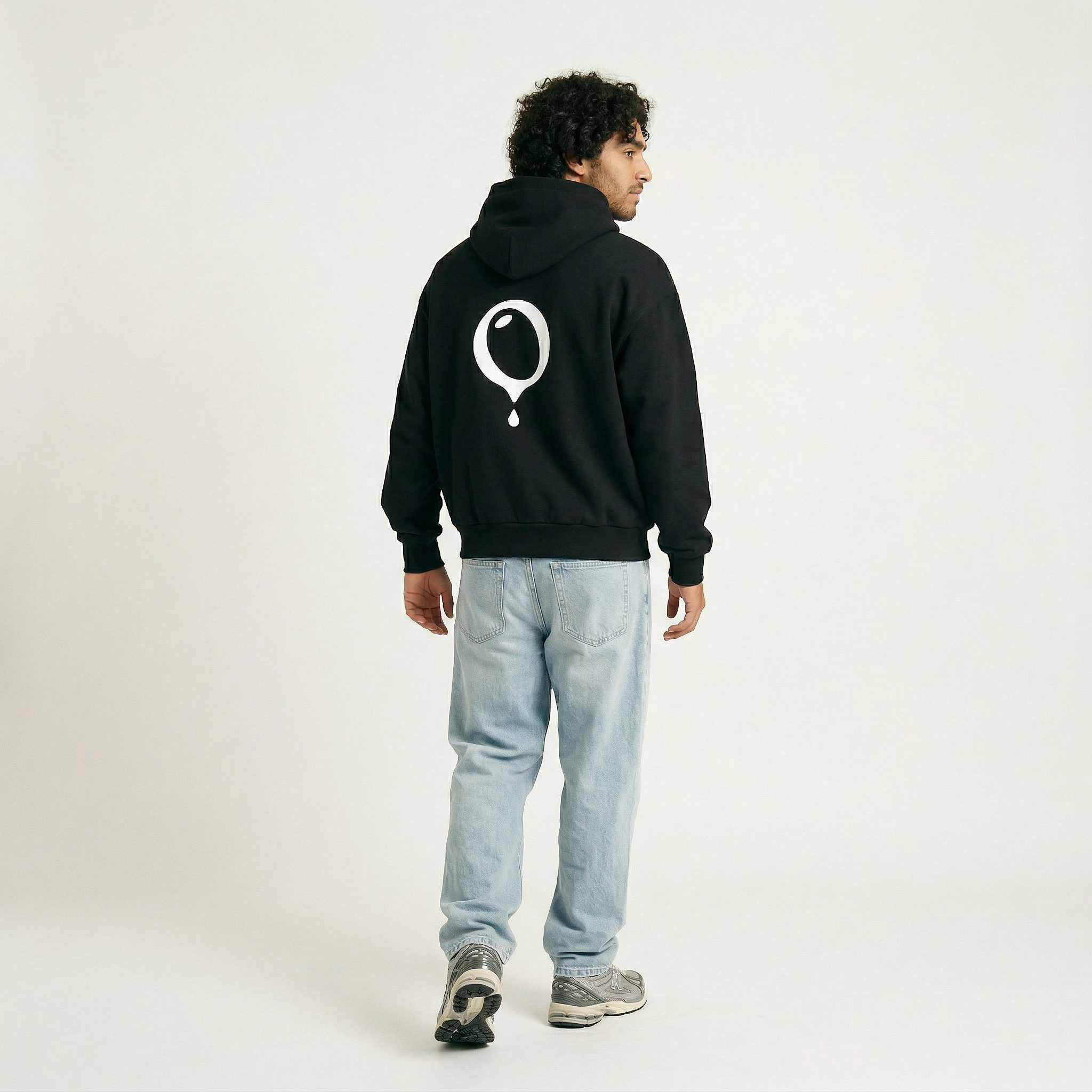 A man stands facing away in the Hoji HOJI HOODIE by Hoji, featuring an oversized fit and a large white balloon graphic on the back, paired with light blue jeans and gray sneakers against a plain light background.