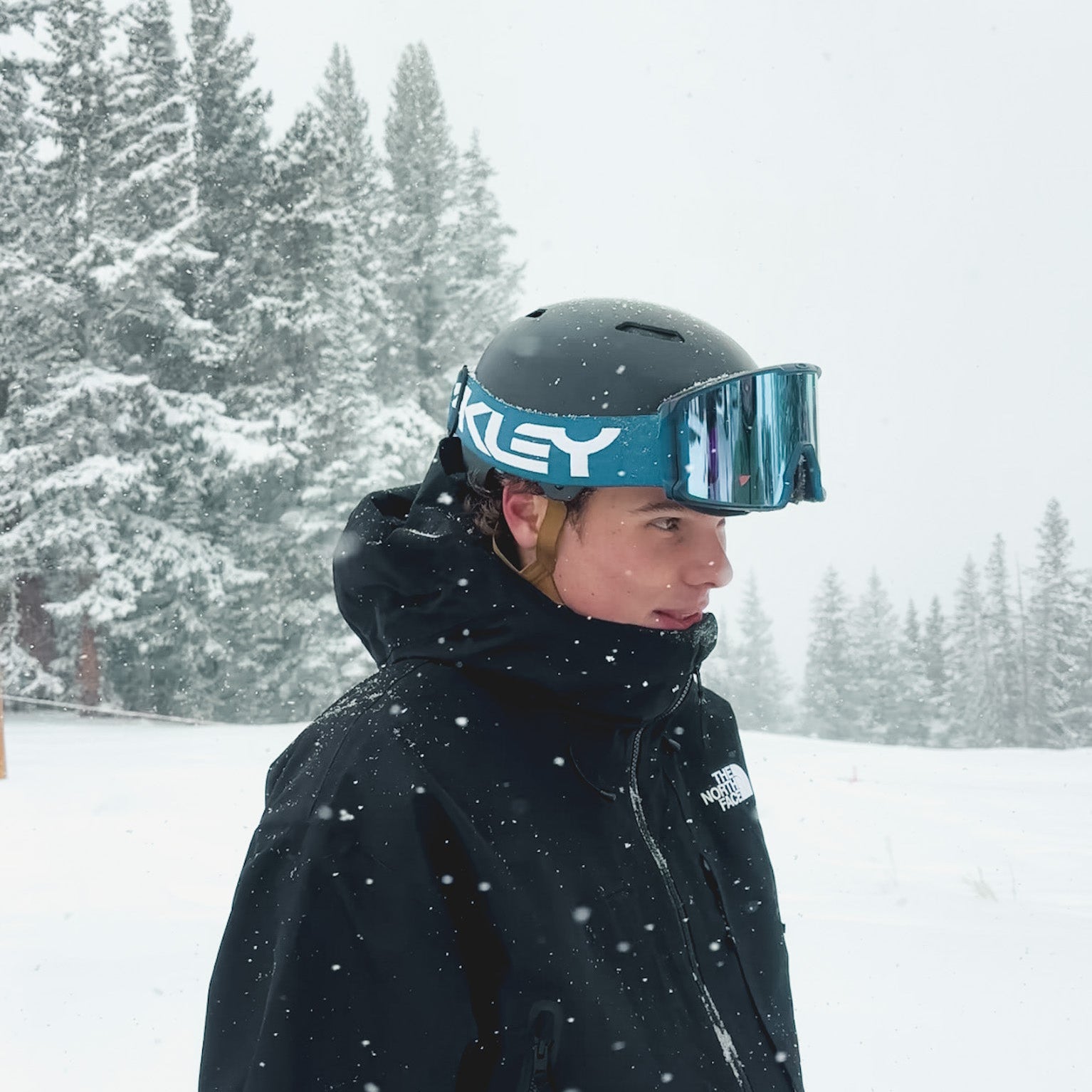 A person in a black jacket and helmet with blue Oakley ski goggles stands in a snowy landscape with snow-covered trees in the background. Snow is falling gently around them.