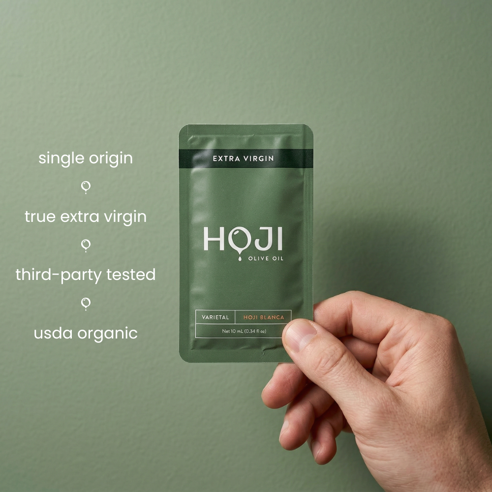 A hand holds a green My Store The 30 Pack olive oil packet labeled extra virgin against a green background. Text on the left highlights single origin, true extra virgin olive oil, third-party tested, and USDA organic.