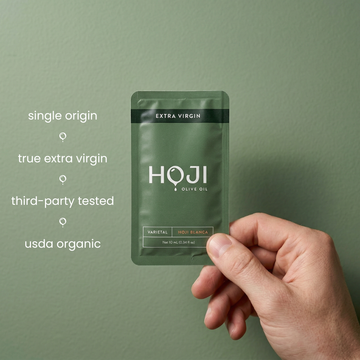 A hand holds a green sachet of My Store 30 PACK (LEGACY) extra virgin olive oil against a matching background. Text on the left: single origin, true extra virgin, third-party tested, USDA organic, and travel-ready olive oil.