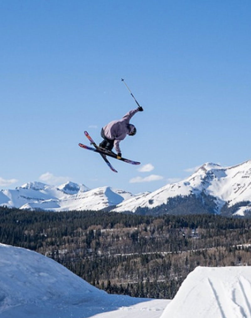 A skier wearing a helmet and goggles performs a mid-air trick above a snowy slope, with snow-covered mountains and trees in the background under a clear blue sky.