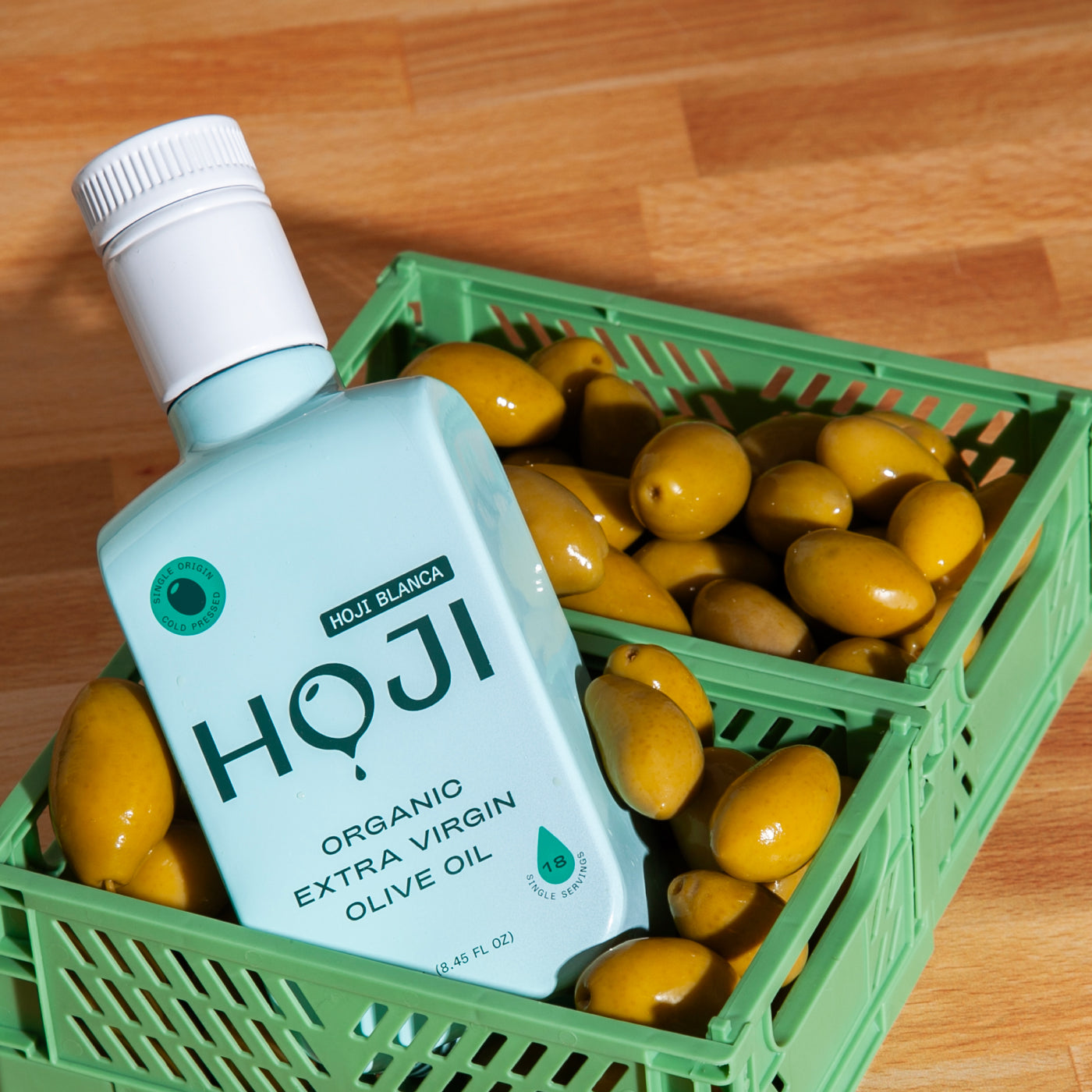 A bottle of HOJI BLANCA BOTTLE by Hoji sits on green crates brimming with green olives, all arranged on a wooden surface.