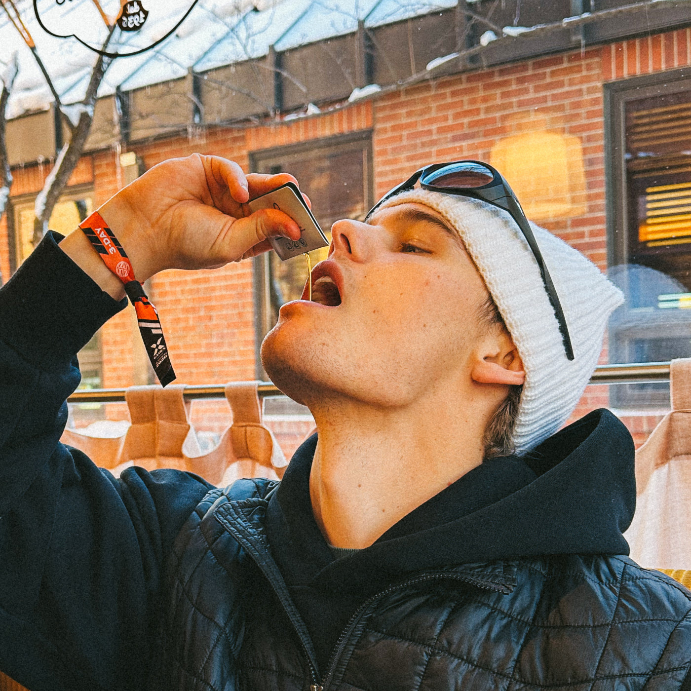 A young person in a white beanie and sunglasses on their head leans back while holding a small metal container above their mouth, appearing to pour something in. A brick building and trees are visible outside the window behind them.