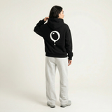 A person stands facing away in a plain light-colored studio, wearing the Hoji HOJI HOODIE—an oversized heavyweight black hoodie with a white circular drip logo on the back—paired with gray sweatpants and gray sneakers.