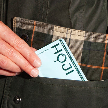 A hand places a light blue HOJI BLANCA 30 PACK olive oil sachet from Hoji—subtle and convenient—into the pocket of a green plaid-lined coat.